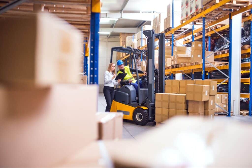 Warehouse workers efficiently managing inventory with a forklift among stacked shipping boxes.